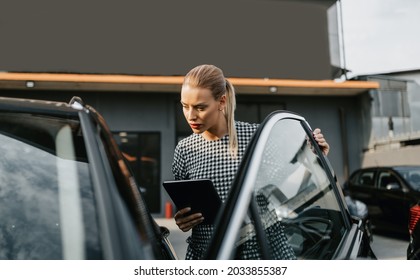 Beautiful Young Woman Working As Used Car Seller. She Is Using Digital Tablet And Checking Car Condition Before Customer Or Buyer. Used Vehicle Dealership.