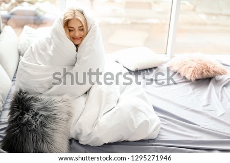 Beautiful young woman wrapped in soft blanket sitting near window at home. Winter atmosphere