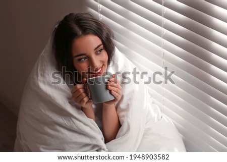 Beautiful young woman wrapped with soft blanket enjoying hot drink near window at home