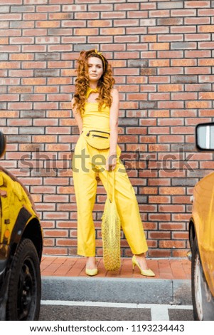 beautiful young woman in yellow clothes with pineapple in string bag standing at parking in front of brick wall