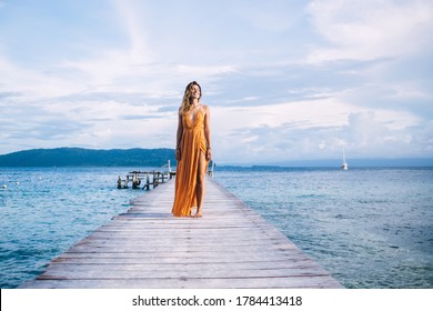 Beautiful young woman in yellow dress smiling and standing on wooden pier against background of sea, mountains and clouds at sunny day - Powered by Shutterstock