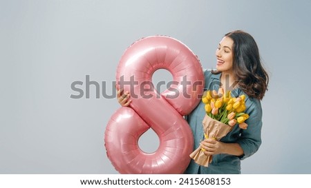 Beautiful young woman with yellow flowers and a balloon in the shape of an eight on gray wall background.
