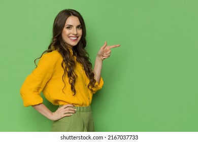 Beautiful young woman in yellow linen shirt is pointing to the side and smiling. Three quarter length studio shot against green background. - Shutterstock ID 2167037733