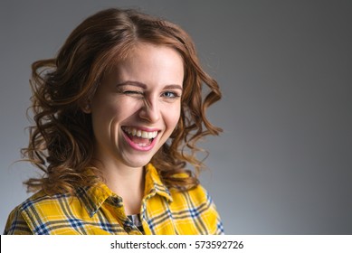 Beautiful Young Woman In A Yellow Shirt Expresses Emotions Wink And Smiling On A Gray Background