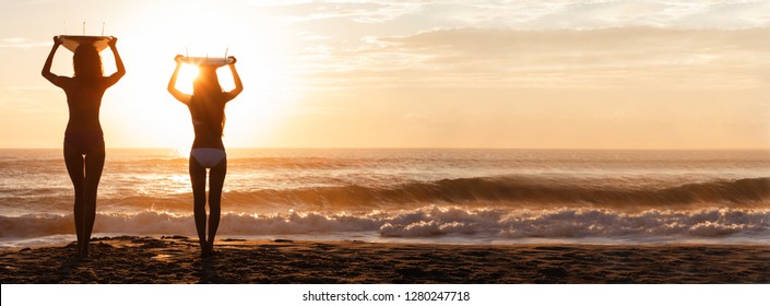 Beautiful young women surfer girls in bikinis with surfboards on a beach at sunset or sunrise panoramic web banner