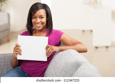 Beautiful youthful African American woman holding blank white sign on grey couch wearing pink shirt.