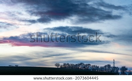 In beautiful,gently rolling arable land,,leafless trees dotted across wintery farmland in southern England,layered clouds glowing red with afterlight beyond.
