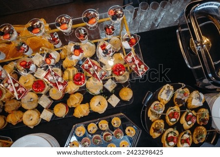 A beautifully arranged catering banquet table showcasing an assortment of delicious snacks and desserts. Guests are enjoying the sumptuous spread in an opulent room with sparkling chandeliers.