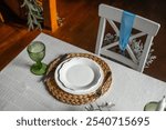 A beautifully arranged dining table features a white plate on a textured placemat, accompanied by green glassware and stylish cutlery, creating an inviting atmosphere.