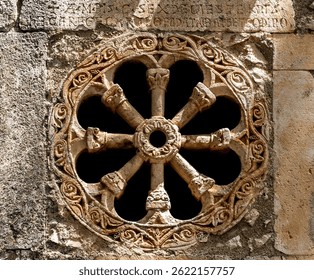 A beautifully carved stone rose window with intricate floral and geometric patterns, set against a weathered stone wall. The design features a circular shape with eight spokes, showcasing medieval cra
