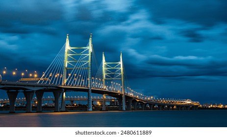 Beautifully illuminated suspension bridge spans across calm river at night, with city lights twinkling in background. Night cable-stayed bridge with street lights under a cloudy sky, long exposure - Powered by Shutterstock