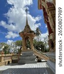 A beautifully ornate golden spire and structure stand proudly at a Thai temple under a bright blue, cloudy sky.