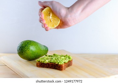 Beautifully Rye Toast Bread With Smashed Green Avocado With Lemon Juice And Black Pepper On Top On A Wooden Table And White Background. Woman Hand Squeezing Half Of Lime On Whole Bread Avocado Toast.