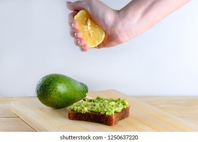 Beautifully Rye Toast Bread With Smashed Green Avocado With Lime Juice And Black Pepper On Top On A Wooden Board And White Background. Woman Hand Squeezing Half Of Lemon On Whole Bread Avocado Toast.