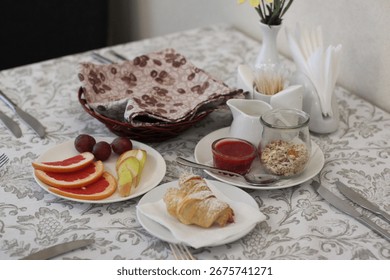 A beautifully set breakfast table featuring a croissant, sliced fruits, jam, and cereals. Includes decorative elements like a basket and flowers.