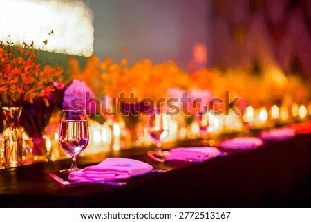 A beautifully set table for a formal wedding reception, gala dinner, or upscale corporate event. A long dining table draped in dark cloth features vibrant floral arrangements with orange blossoms and magenta pink flowers running down the center. Each place setting includes a light pink napkin, polished silverware, and a stemmed water glass reflecting ambient light. Small votive candles scattered among the flowers cast a soft golden glow. Warm overhead lighting and shallow depth of field create an intimate celebratory mood with soft bokeh effect.