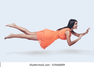 Beauty In The Air. Full Length Studio Shot Of Attractive Young Woman In Orange Dress Hovering In Air And Smiling