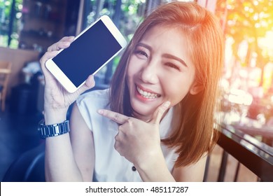 beauty asian girl smiling and showing smartphone with empty screen for your text  in cafe background