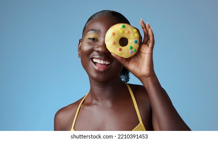 Beauty, black woman and portrait of donut on face with blue studio wall for happy summer style. Matching, beautiful and fun african american girl with yellow makeup for quirky fashion campaign.