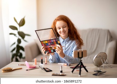 Beauty Blog. Young beautiful teen doing makeup, recording cosmetics product review, showing her eyeshadow palette to camera. Girl talking in front of phone on tripod, filming video for her channel