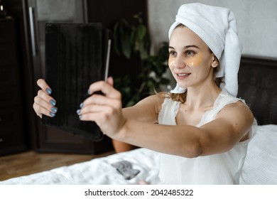 Beauty blogger, vlogger, influencer. Young Woman in pajamas, towel on her head and with cosmetic collagen patches under eyes recording video clip on tablet. Spa, skincare and wellness - Powered by Shutterstock