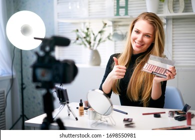 Beauty blogger woman filming daily make-up routine tutorial near camera. Influencer girl live streaming cosmetics product review. Vlogger female recommends eye shadow palette showing thump up sign.