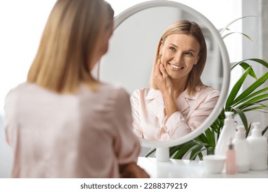 Beauty Care. Attractive Middle Aged Woman Looking In Mirror At Home And Smiling To Her Reflection, Beautiful Mature Blonde Lady Touching Face, Enjoying Flawless Skin After Anti-Aging Treatments