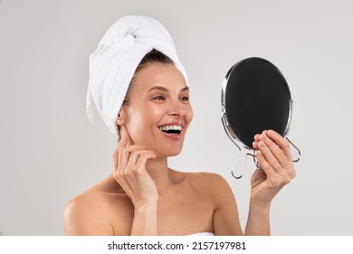Beauty Care Concept. Attractive woman with towel on head holding magnifying mirror, happy smiling female looking at reflection, enjoying her perfect skin while posing over gray background, copy space