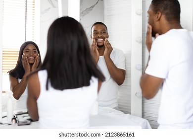 Beauty Care Concept. Happy Black Couple Making Morning Routine In Bathroom, Millennial African American Spouses Applying Face Cream And Smiling To Their Reflection In Mirror, Selective Focus