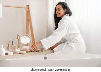 Beauty Care. Happy Lady Caring For Herself Using Cosmetics Products Sitting On Bathtub In Modern Bathroom At Home, Wearing Bathrobe. Wellness And Spa, Bodycare Concept