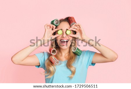 Beauty care at home concept. Laughing young girl in curlers puts pieces of cucumber on eyes