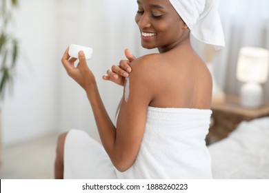 Beauty Care Product, Dry Female Skin And Hydration. Happy Cute Millennial African American Woman In White Towel After Shower Applies Cream To Body And Holds Jar With Empty Space, In Bedroom Interior