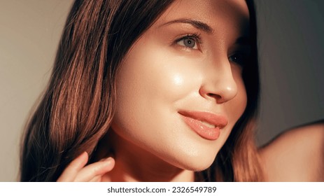 Beauty care. Radiant face. Moisturized skin. Cosmetology treatment. Pretty smiling young fresh glowing woman with glossy lip makeup enjoying sunshine isolated on beige.