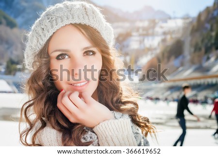 Beauty caucasian woman going to ice skating outdoor. She dressed in white winter pullover and warm hat. Healthy lifestyle and sport concept at olympic rink, mountain landscape, snowy blizzard