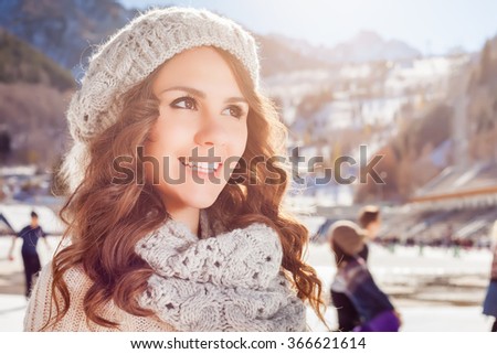 Beauty caucasian woman going to ice skating outdoor. She dressed in white winter pullover and warm hat. Healthy lifestyle and sport concept at olympic rink, mountain landscape, snowy blizzard