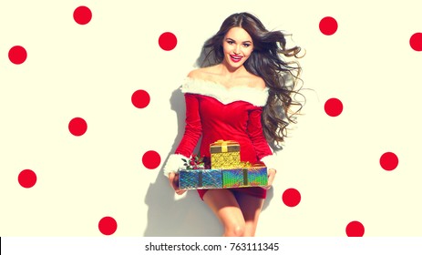 Beauty Christmas Fashion Model Girl With Xmas Gift Boxes, Wearing Red Santa Dress Holding Gifts. Sexy Brunette Young Woman Portrait. Stylish Female, Fashionable Party Clothes And Accessories, Makeup