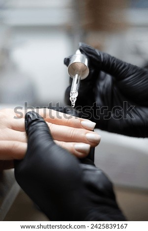 Beauty concept. A manicurist in black latex gloves makes a hygienic manicure and paints the client's nails with gel polish and drips oil on the cuticles in a beauty salon. Close-up. Vertical.