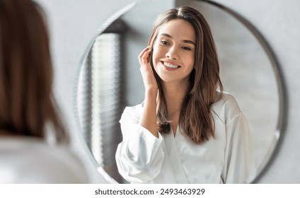 Beauty Concept. Portrait Of Attractive Happy Woman Looking At Mirror In Bathroom, Beautiful Millennial Lady Wearing White Silk Robe Smiling To Reflection, Enjoying Her Appearance, Selective Focus