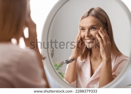 Beauty Concept. Portrait Of Attractive Mature Woman Looking At Mirror In Bathroom, Beautiful Middle Aged Lady Touching Her Flawless Face Skin And Smiling To Reflection, Selective Focus