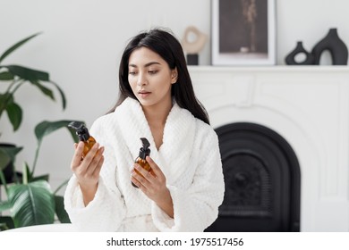 Beauty concept. Thoughtful young asian woman holding glass bottles in hands, choose skincare and body care organic cosmetics. Female in white bathrobe spending morning in modern bathroom
