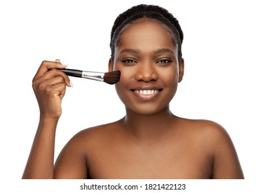 beauty, cosmetics and people concept - portrait of happy smiling young african american woman with make up brush applying blush to her face over white background