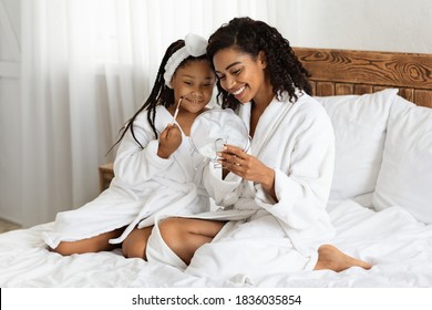 Beauty Day With Mom. Black Mother And Her Daughter In Bathrobes Applying Makeup Together, Young Woman Holding Magnifying Mirror And Little Girl Playing With Brush, Sitting Together On Bed At Home