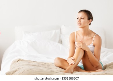 Beauty, Depilation, Epilation And Bodycare Concept - Beautiful Woman Touching Her Bare Legs Sitting On Bed At Home Bedroom