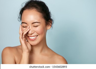 Schönes Gesicht. Lächelnde asiatische Frau, die gesundes Hautporträt berührt. Schönes, fröhliches Mädchenmodell mit frisch glühender, hydrierter Gesichtshaut und natürlichem Make-up auf blauem Hintergrund im Studio. Hautpflegekonzept