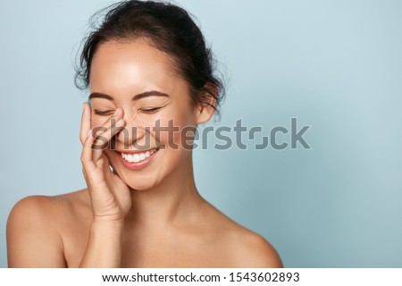 Beauty face. Smiling asian woman touching healthy skin portrait. Beautiful happy girl model with fresh glowing hydrated facial skin and natural makeup on blue background at studio. Skin care concept