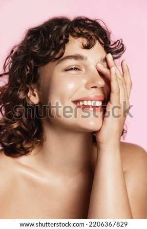 Beauty face. Smiling curly teen girl touching healthy skin portrait. Beautiful happy woman model with fresh glowing hydrated facial skin and natural makeup on pink background at studio.