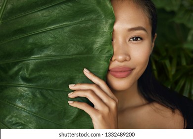 Beauty face. Woman model with natural makeup and healthy skin behind green leaf plant. Portrait of beautiful asian girl with nude nails, big lips and sexy smile in tropical nature