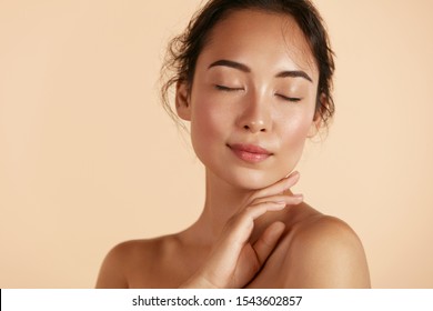 Beauty face. Woman with natural makeup and healthy skin portrait. Beautiful asian girl model touching fresh glowing hydrated facial skin on beige background closeup. Skin care concept