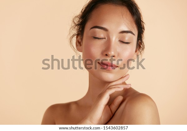 Beauty face. Woman with natural makeup and\
healthy skin portrait. Beautiful asian girl model touching fresh\
glowing hydrated facial skin on beige background closeup. Skin care\
concept