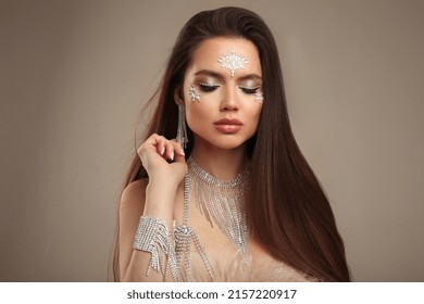 Beauty Fashion Art Glamour Girl Portrait With Gems On Face. Beautiful Brunette Woman In Diamond Jewelry Accessories Isolated On Beige Studio Background.